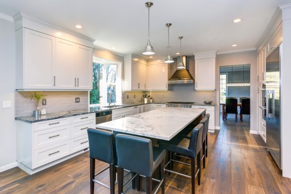 Luxury home interior boasts Beautiful black and white kitchen with custom white shaker cabinets, endless marble topped kitchen island with black leather stools over wide planked hardwood floor.