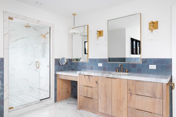 A luxurious bathroom with a wood vanity cabinet, blue tiles on the walls, gold accented lights and mirrors, and a large walk-in shower lined with marble tiles. No brands or labels.
